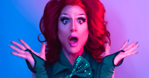 Primer plano de una artista drag con una expresión de gran sorpresa y la boca abierta. Lleva una peluca de cabello rojizo voluminoso, un maquillaje de ojos muy elaborado con pestañas largas y dramáticas, guantes verdes y un lazo a juego. El fondo está iluminado con luces de neón en tonos azules y rosados.