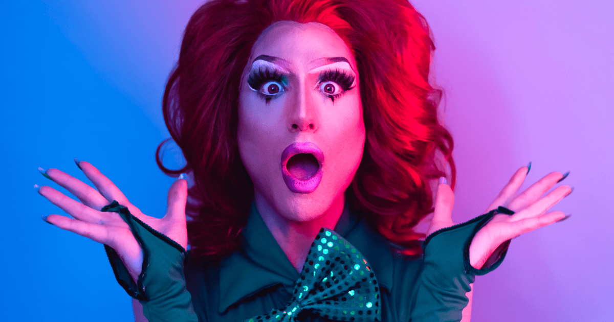 Primer plano de una artista drag con una expresión de gran sorpresa y la boca abierta. Lleva una peluca de cabello rojizo voluminoso, un maquillaje de ojos muy elaborado con pestañas largas y dramáticas, guantes verdes y un lazo a juego. El fondo está iluminado con luces de neón en tonos azules y rosados.