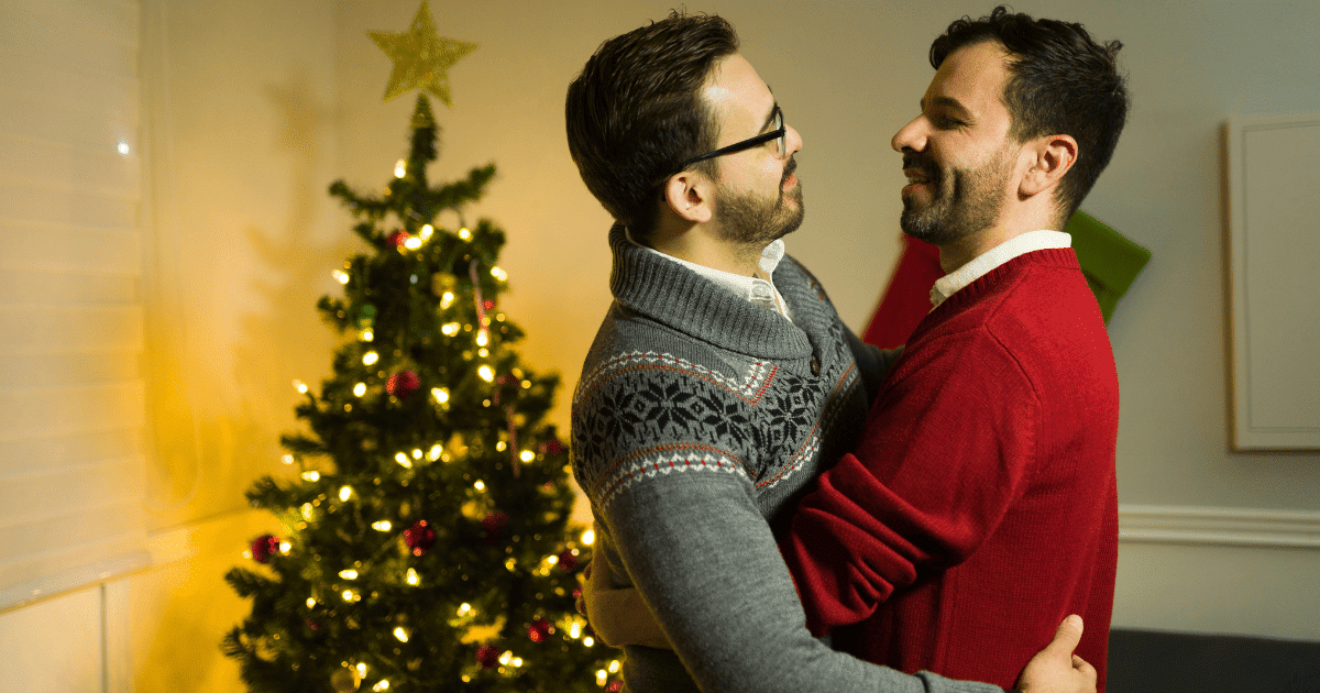 Una pareja de hombres jóvenes abrazándose cariñosamente frente a un árbol de Navidad iluminado en un salón acogedor.