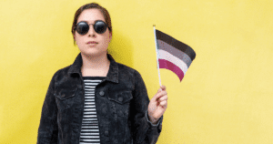 Mujer joven con gafas de sol sosteniendo la bandera del orgullo asexual (franjas negra, gris, blanca y morada) sobre un fondo amarillo.