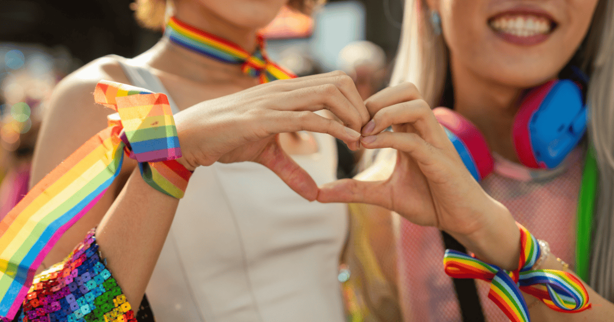 Dos personas en Andalucía haciendo el símbolo de un corazón con sus manos, adornadas con lazos y pulseras arcoíris durante la celebración del Orgullo LGTBI+.