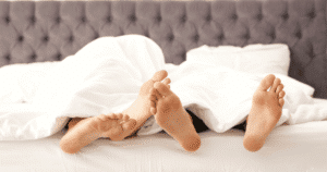Primer plano de varios pares de pies asomando por debajo de una sábana blanca en una cama con cabecero gris.